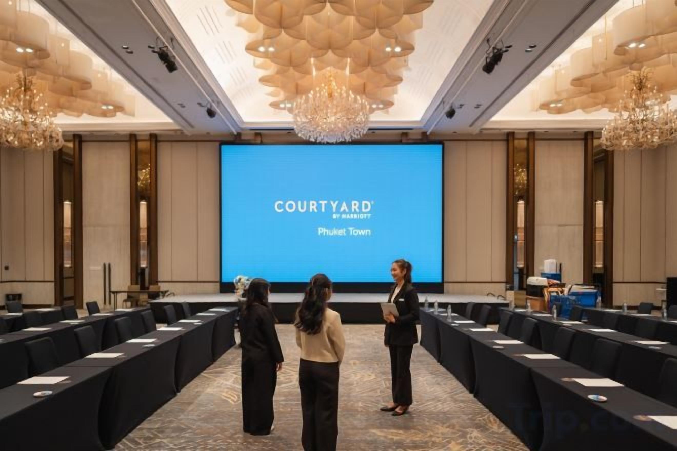 Uma sala de conferências com uma grande tela exibindo 'COURTYARD by Marriott Phuket Town', uma mesa comprida com cadeiras e um lustre.