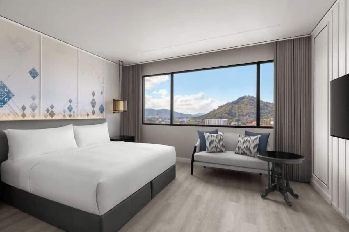 Habitació d'hotel moderna amb un llit gran, un sofà, una taula petita i una finestra ampla amb vista a la muntanya.
