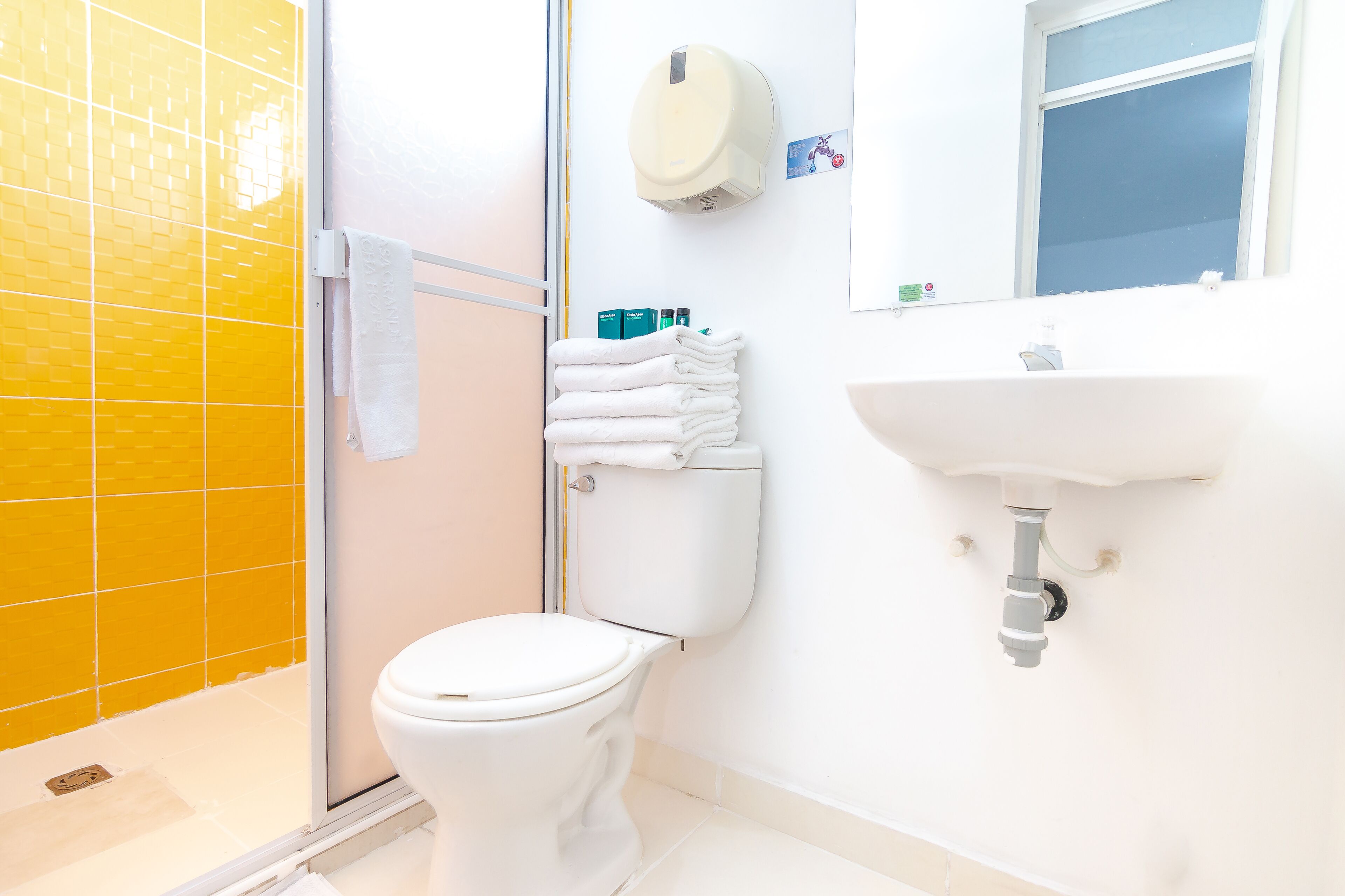 A bathroom with a yellow-tiled shower, a white toilet, a sink, and a towel rack with folded towels.