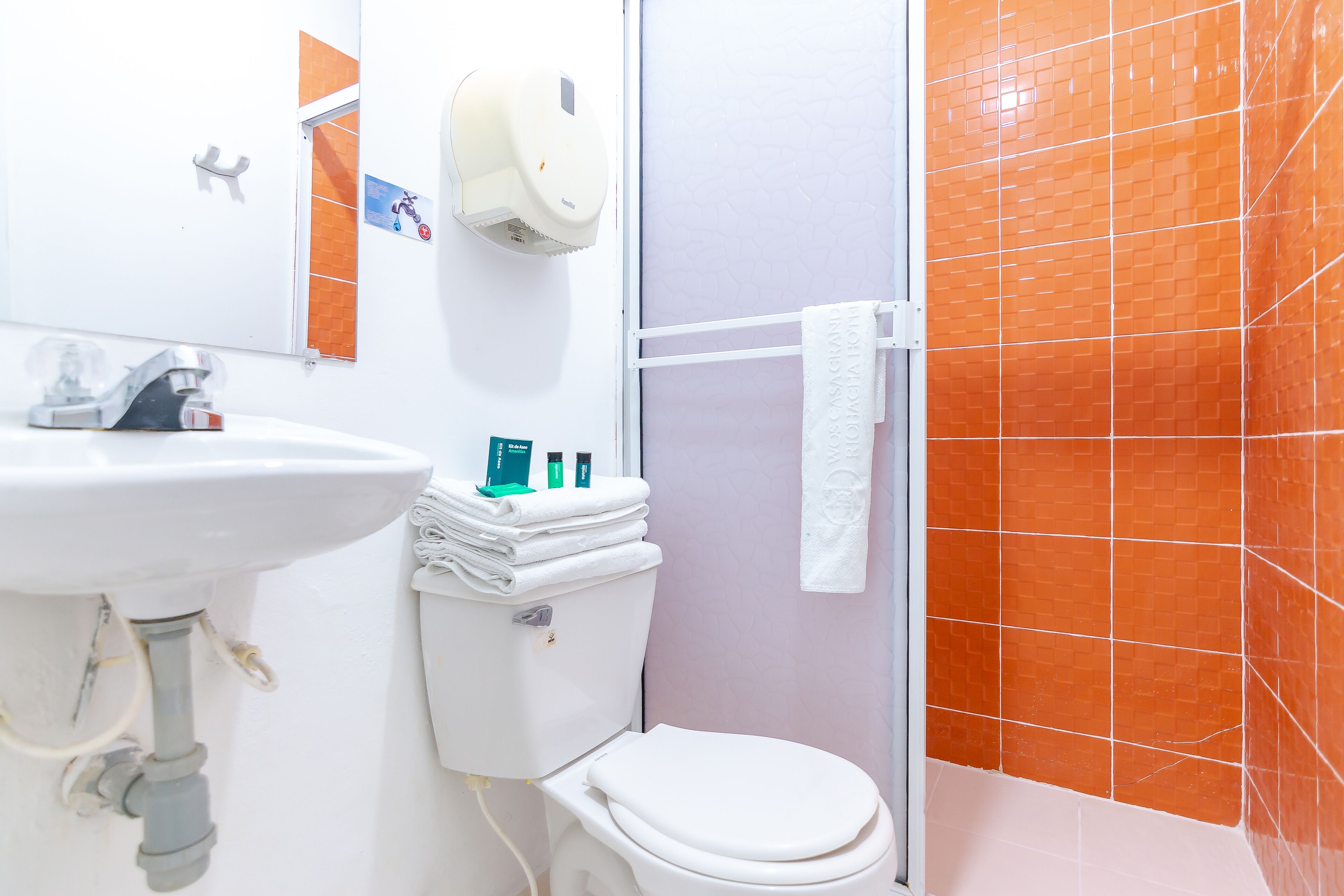Une salle de bain avec un lavabo, des toilettes et une douche, le tout de couleur blanche. On y trouve un porte-serviettes sur lequel est accrochée une serviette blanche.