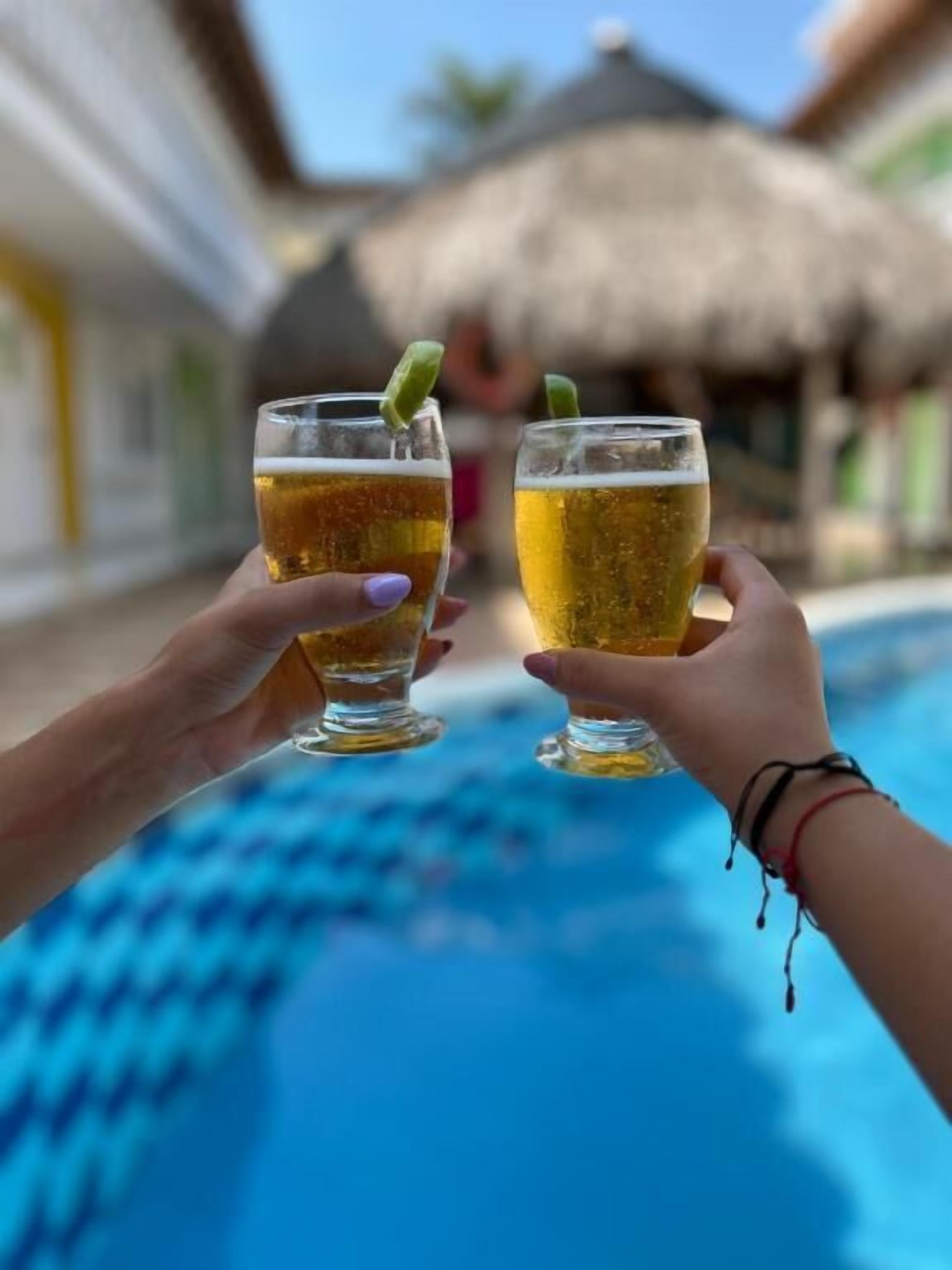 Dos manos sosteniendo vasos de cerveza junto a una piscina.