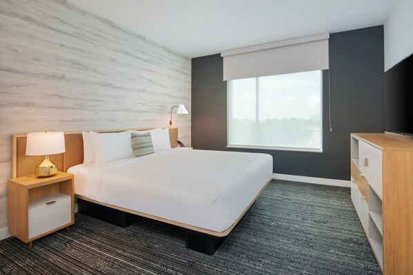 A hotel room with a large bed, a nightstand with a lamp, a dresser, and a window with a view of the city.