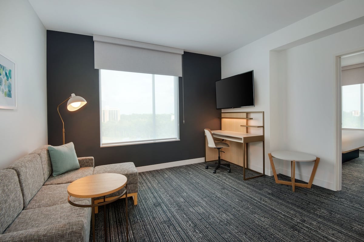 Una habitación de hotel moderna con un sofá gris, una mesa redonda de madera, un escritorio con una silla, un televisor de pantalla plana y un ventanal con vistas.
