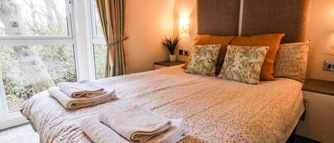 A neatly made bed with floral bedding and two rolled towels, a nightstand with a lamp, and a window with curtains.