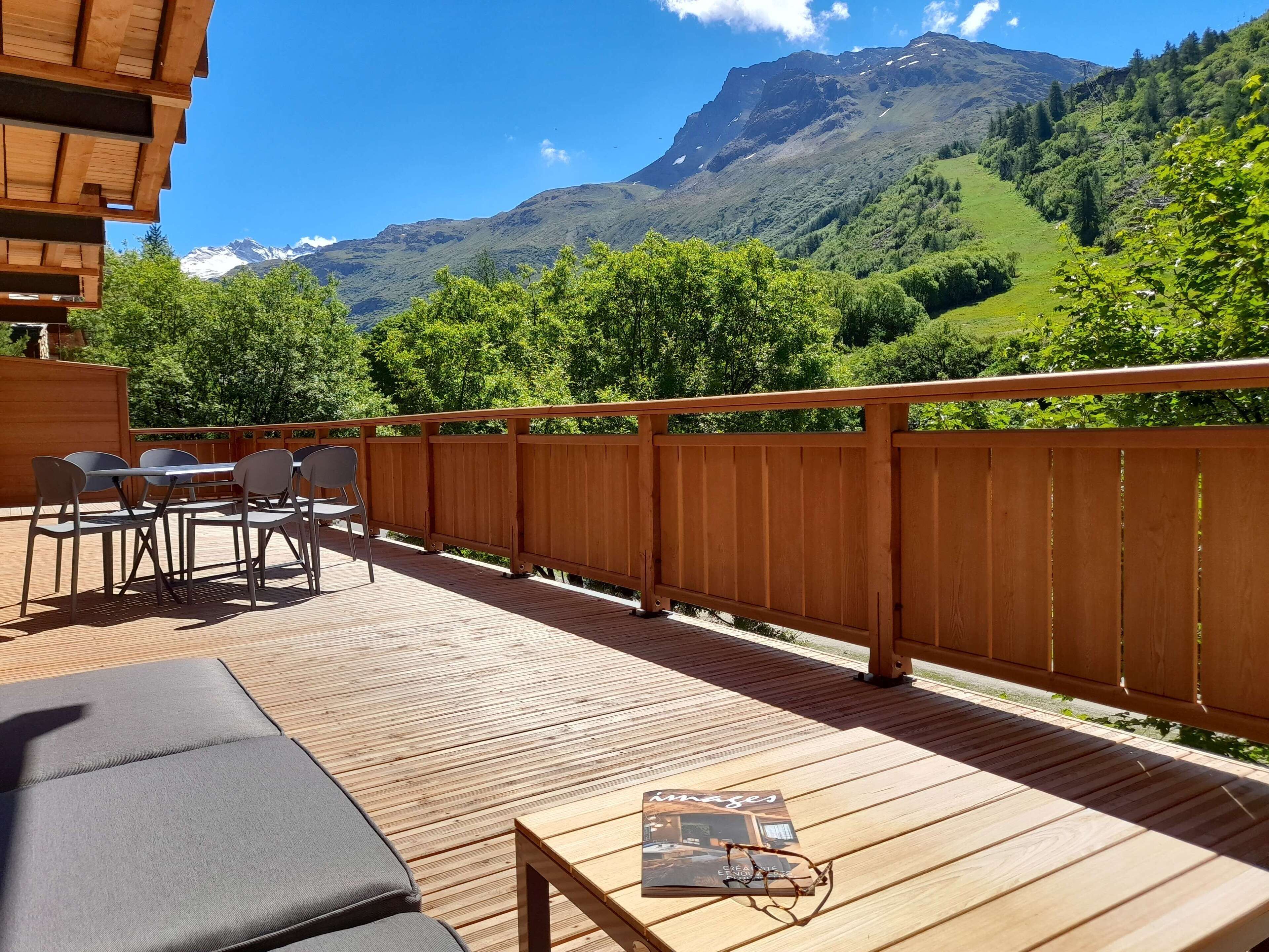 Un terrazzo in legno con posti a sedere all'aperto, un tavolo con una rivista e dei bicchieri, e vista sulle montagne.