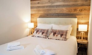 Una cama bien tendida con sábanas blancas y dos almohadones decorativos, un cabecero de madera y dos lámparas de noche sobre mesitas de madera.