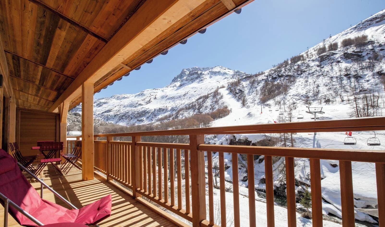 Une terrasse en bois, agrémentée de chaises et de tables, qui offre une vue sur un paysage montagneux enneigé, avec des remontées mécaniques.