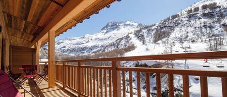 Terraza de madera con sillas y mesas, con vistas a un paisaje montañoso nevado y telesillas.
