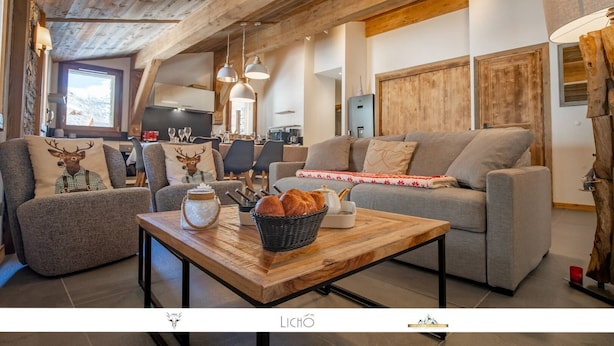 Un salon chaleureux avec un canapé, des fauteuils, une table basse en bois et une cuisine en arrière-plan.