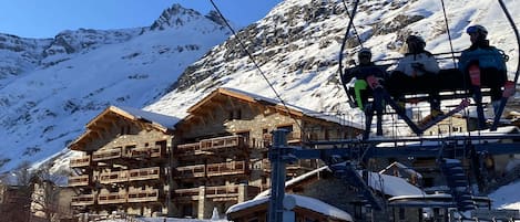 A ski lift with skiers, a wooden chalet with balconies, and a mountainous backdrop.