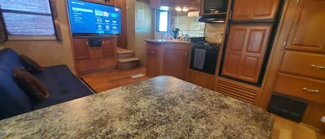 A kitchen with granite countertops, wooden cabinets, a built-in oven, and a flat-screen TV mounted on the wall.
