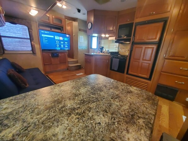 A kitchen with granite countertops, wooden cabinets, a built-in oven, and a flat-screen TV mounted on the wall. - Hilton's Hometown Tower-view Hot-tub Retreat (Cisco)