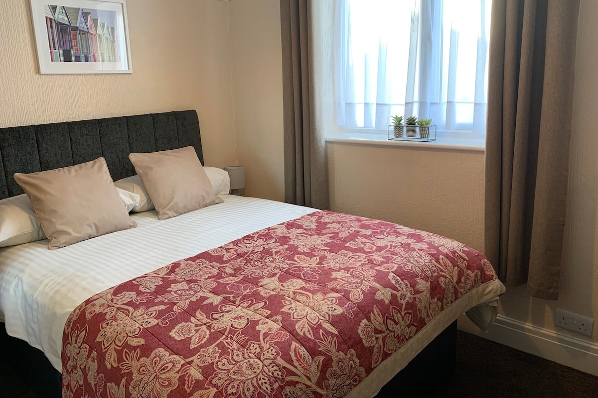 A hotel room with a bed, a headboard, a window with curtains, and a framed picture on the wall.