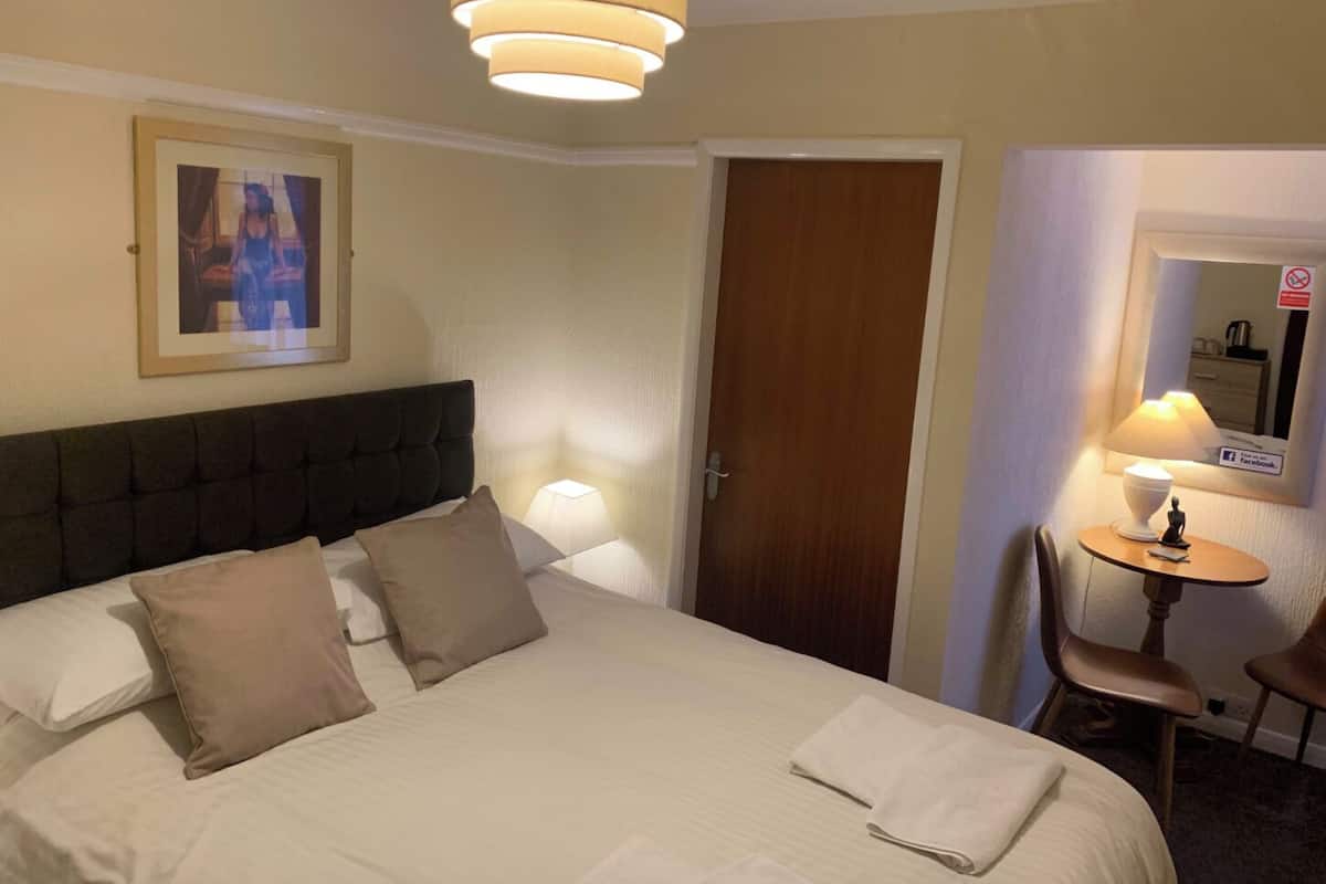 A hotel room with a large bed, a headboard, a bedside table with a lamp, a small table with a chair, and a door.