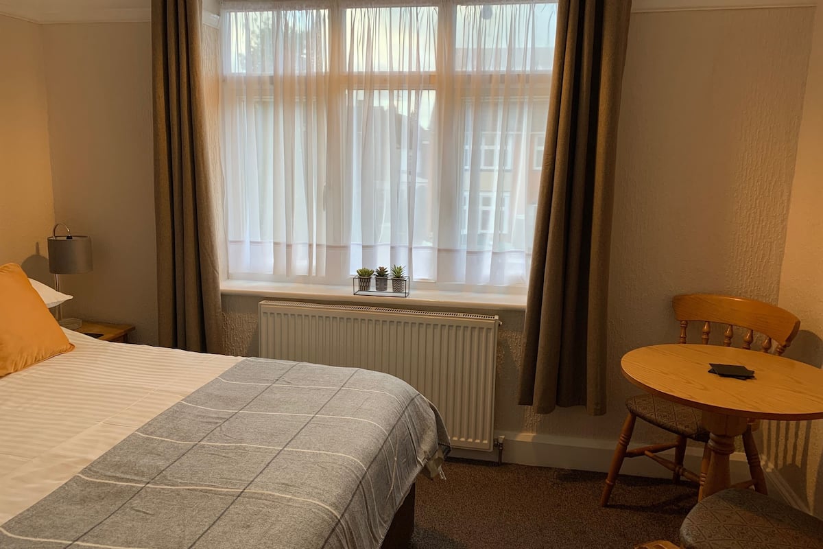 A hotel room with a bed, a table, a chair, a window with sheer curtains, a radiator, and a lamp on a bedside table.