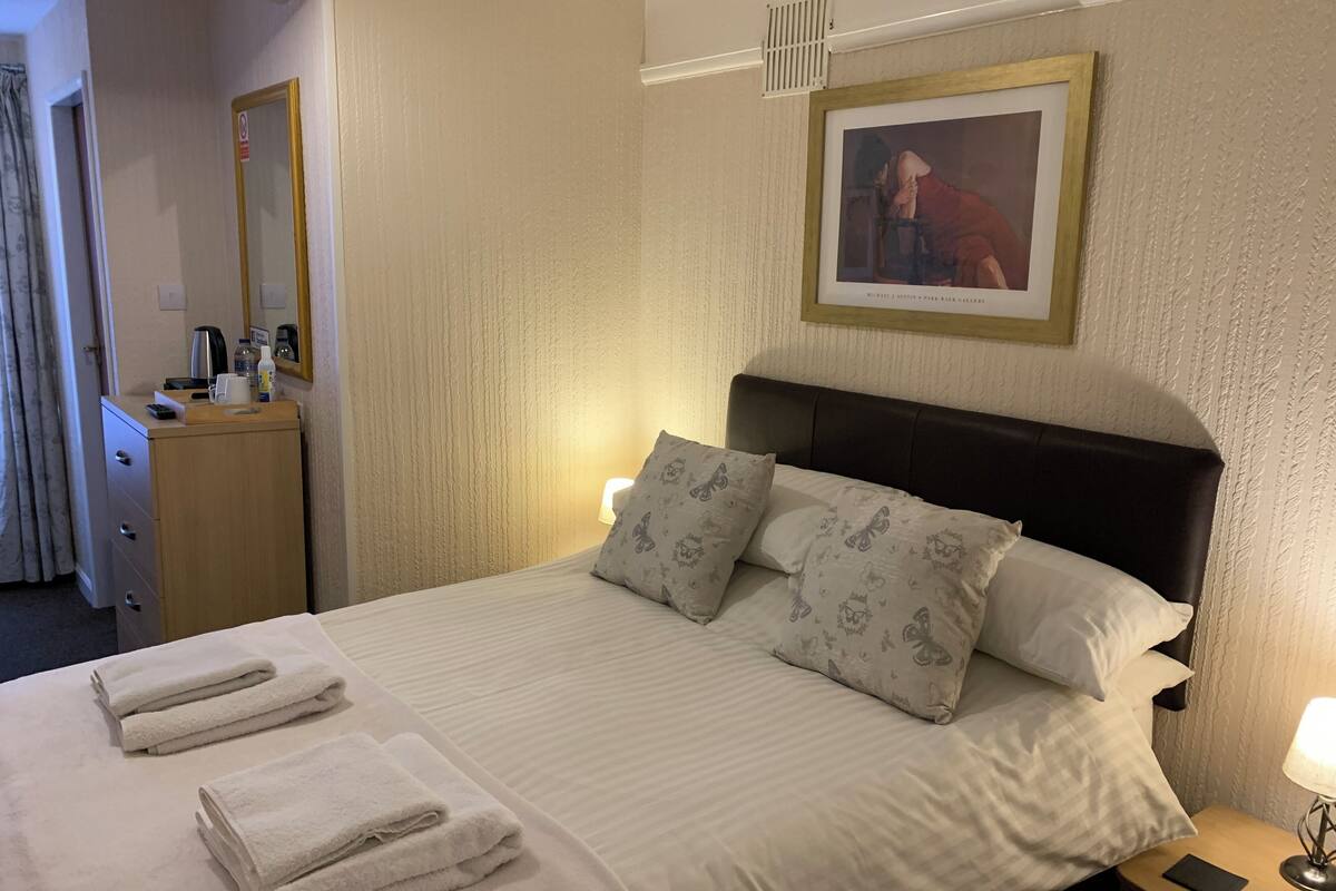 A neatly made bed with pillows and folded towels, a bedside table with a lamp, a framed picture on the wall, and a dresser with a mirror and toiletries.