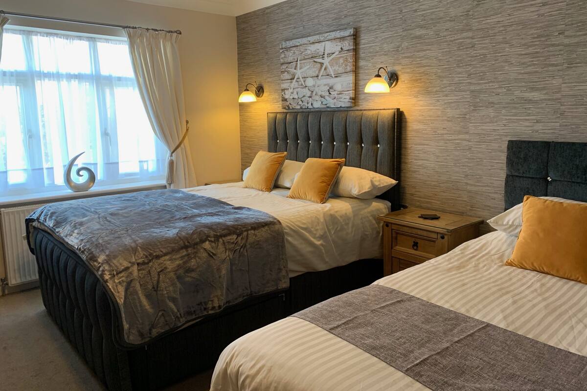 A hotel room with two beds, a large window with curtains, a decorative wall piece, and two wall-mounted lamps.