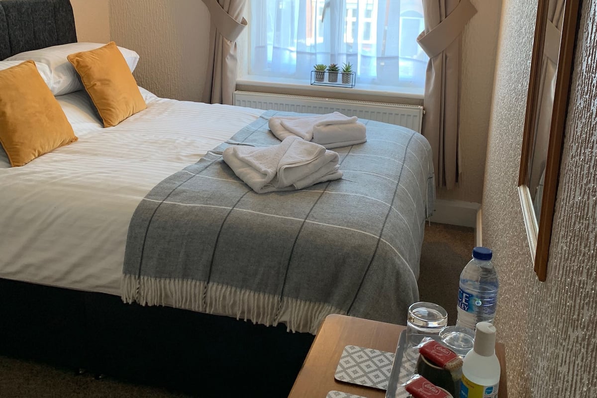 A neatly made bed with a grey blanket, two white towels, and two mustard-colored pillows. There is a small table with a bottle of water, a glass, and a tissue box. A window with sheer curtains and a view of buildings is visible.
