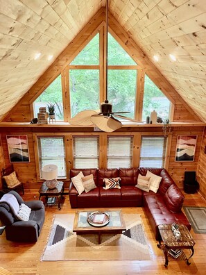 Een gezellig chalet interieur met een slaapkamer op een entresol, houten wanden, een bruine leren bank en een groot raam met uitzicht op groen.