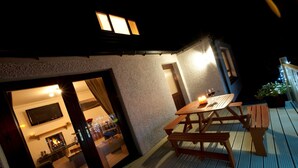 An outdoor patio with a wooden table and benches, a TV mounted on the wall, and a view into the house through a glass door. - Abhainn Cottage (Isle of Lewis)