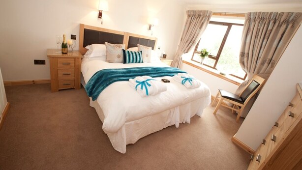 A bedroom with a large bed, bedside tables, a window with curtains, and a chair. - Abhainn Cottage (Isle of Lewis)