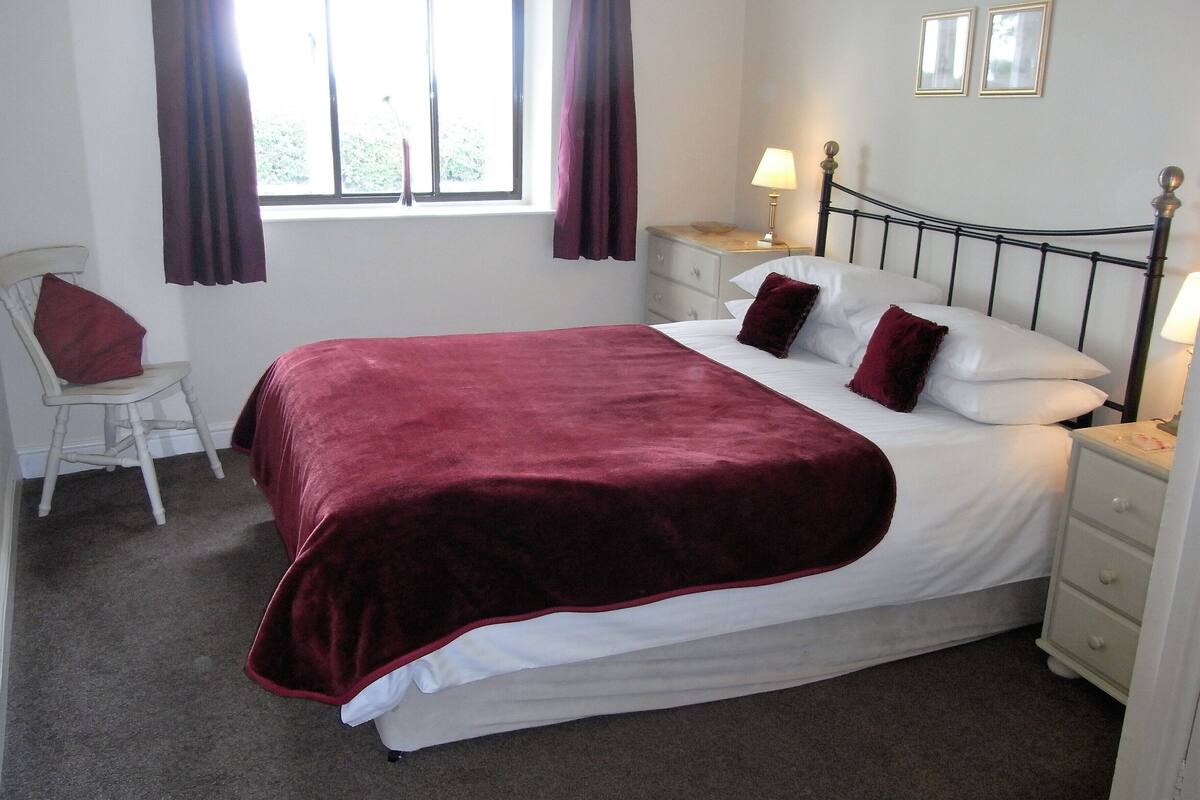 A neatly made bed with a red blanket, white pillows, and a bedside lamp. There is a chair with a red cushion, a window with curtains, and framed pictures on the wall.