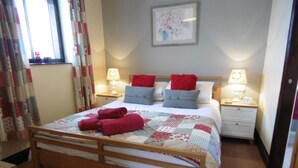 A neatly made bed with red and blue pillows, a quilt, and two red towels. There are two bedside tables with lamps, a framed picture on the wall, and a window with curtains.