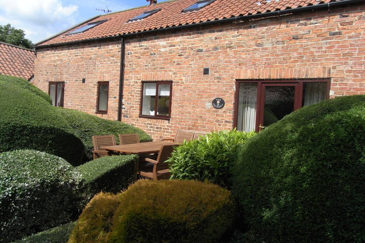 A brick building with a tiled roof, a sign reading "The Oranary," and a patio area with a table and chairs.
