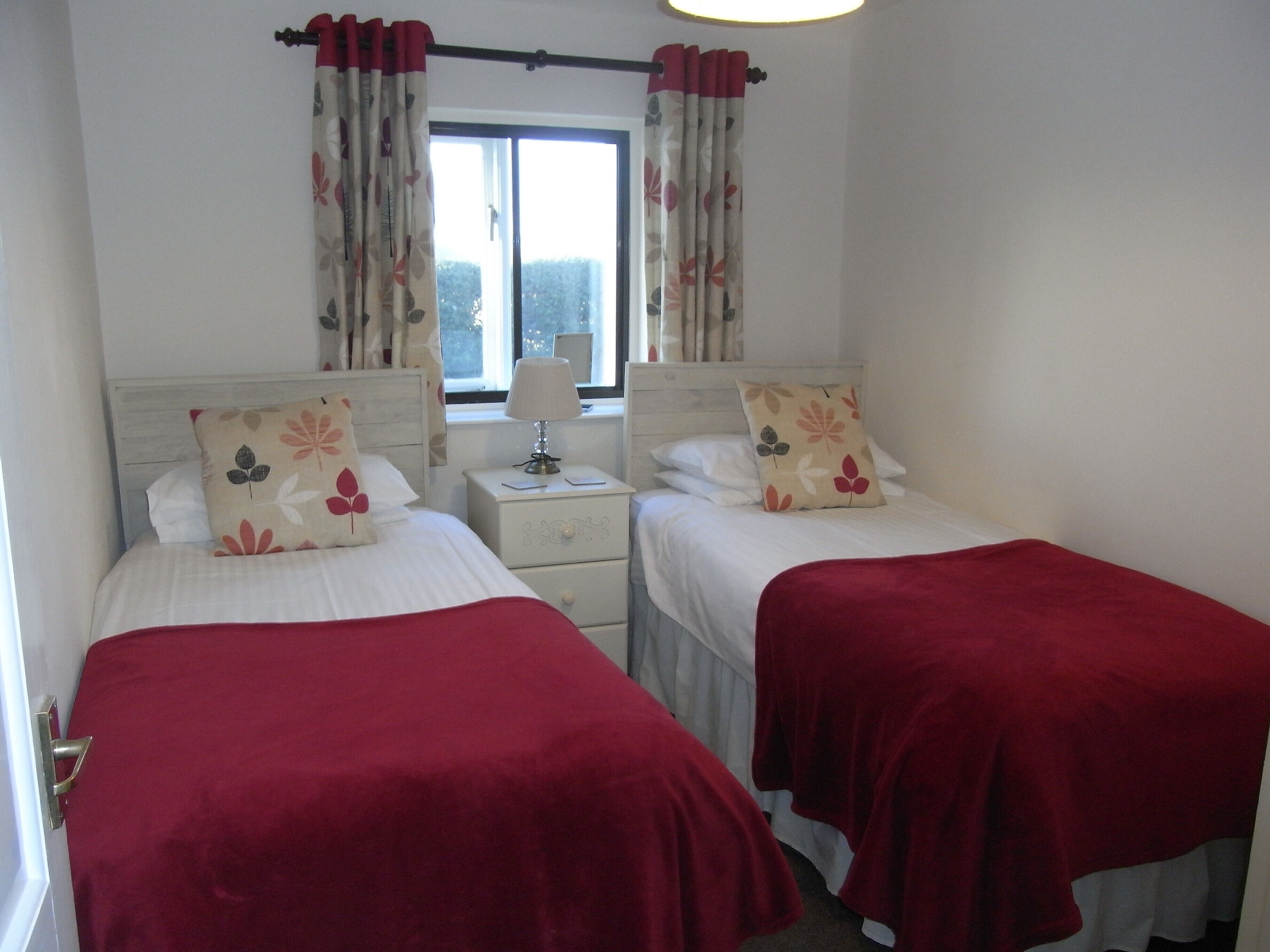 Une chambre double avec deux lits, chacun recouvert d’une couverture rouge, avec une literie blanche et des coussins à motifs floraux assortis. Il y a une table de chevet avec une lampe entre les deux lits, ainsi qu’une fenêtre avec des rideaux.