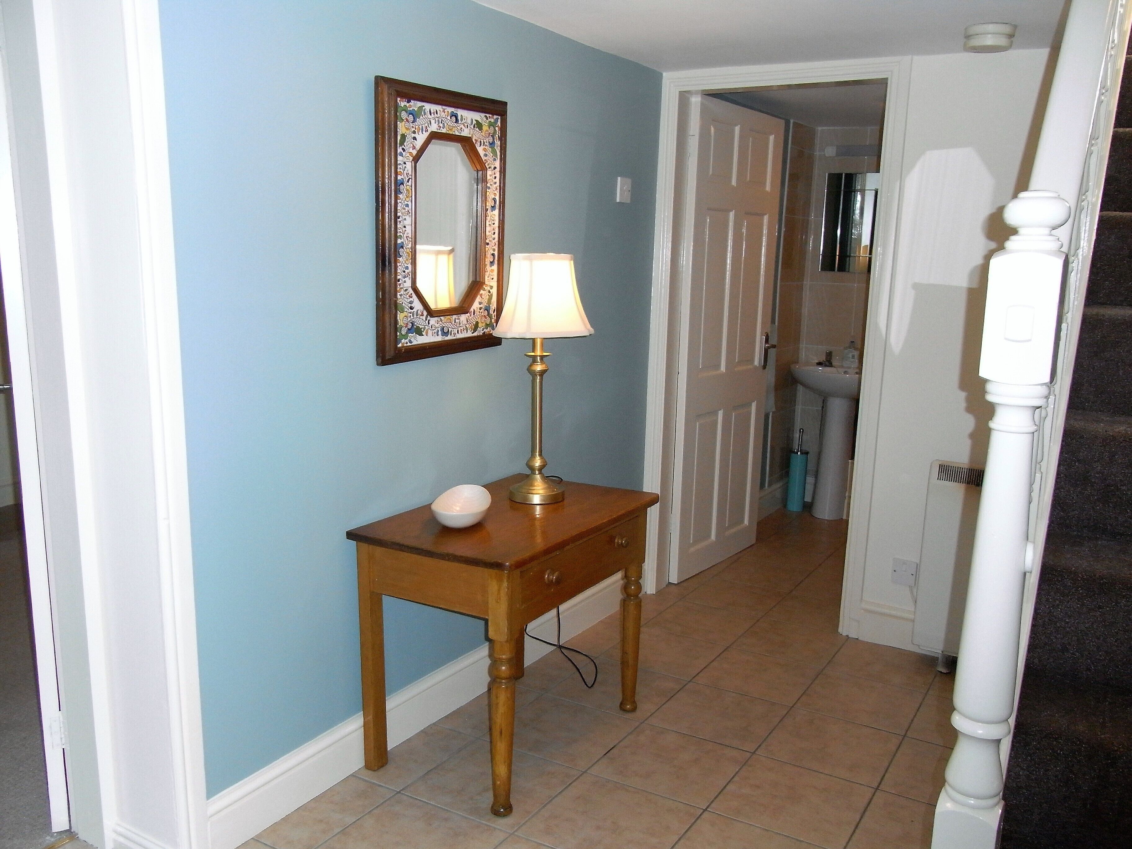A hallway with a wooden console table, a mirror, and a lamp. A door leads to a bathroom.