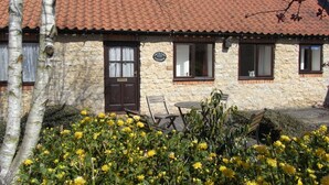 A stone cottage with a tiled roof, a wooden door, and windows with curtains. There is a bench and a table outside.