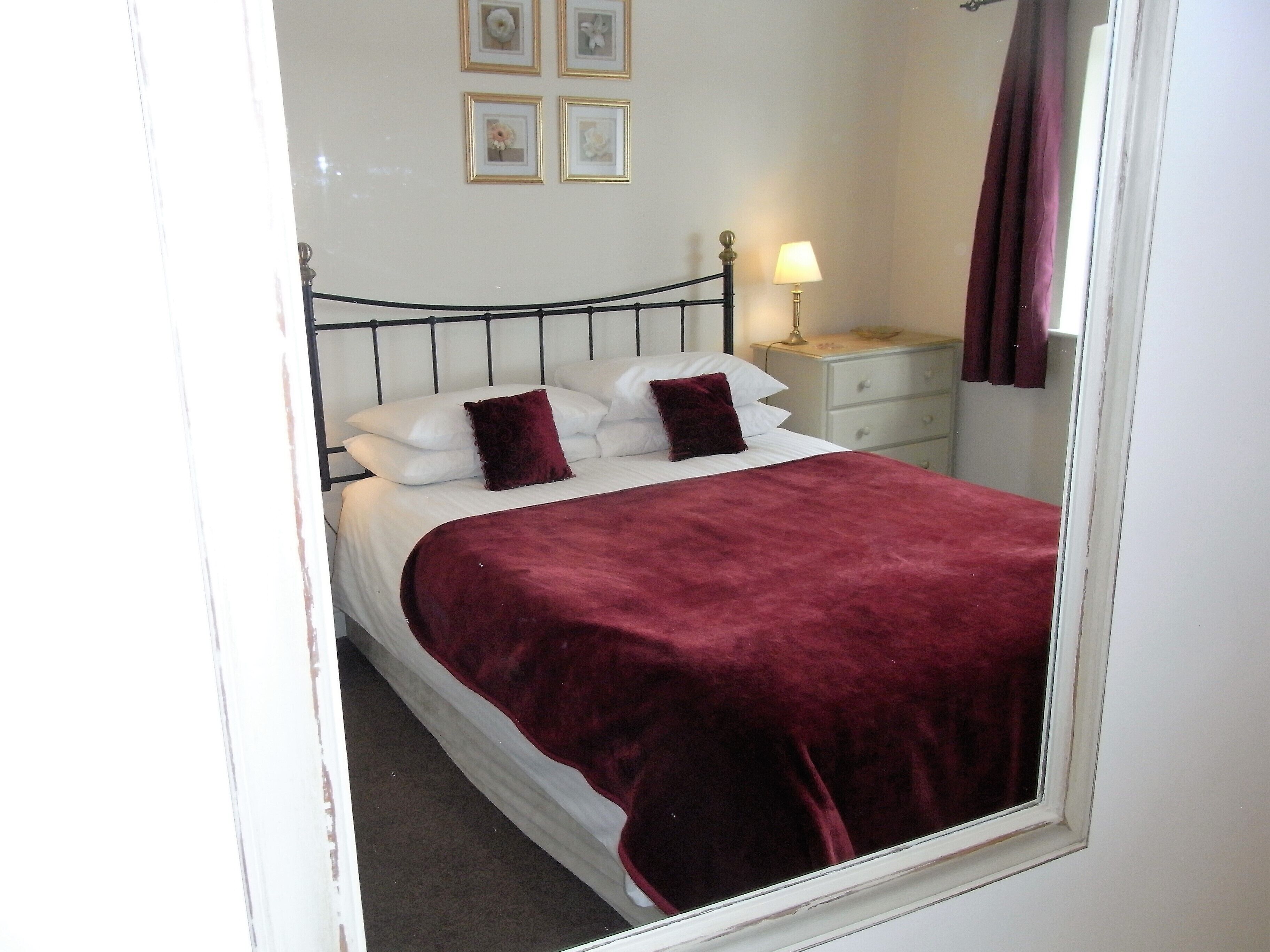 A neatly made bed with a red blanket, white pillows, and a black metal headboard. There is a bedside table with a lamp and a framed picture on the wall above the bed.