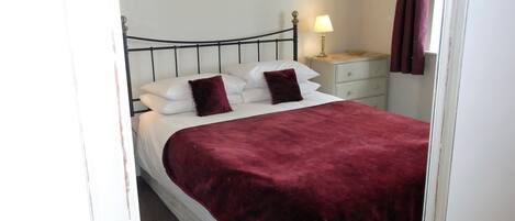 A neatly made bed with a red blanket, white pillows, and a black metal headboard. There is a bedside table with a lamp and a framed picture on the wall above the bed.