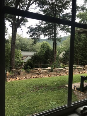 A view through a window of a backyard with a stone wall, a grassy lawn, and a wooden deck. - Sunset Ridge (Asheville)