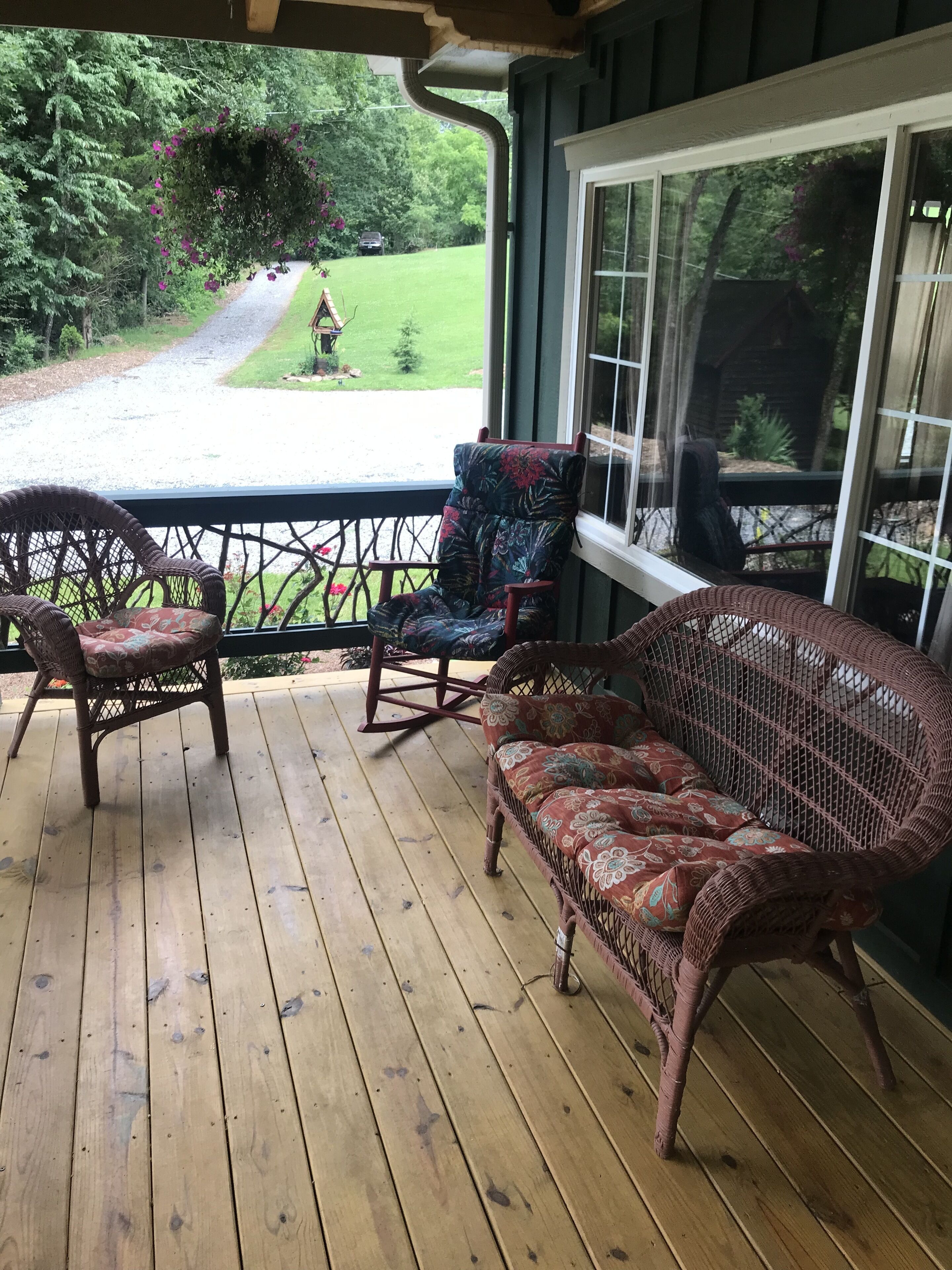 Eine Holzterrasse mit Korbmöbeln, darunter eine Couch und Stühle, mit Blick auf eine Rasenfläche mit einem Pfad und einem kleinen Gebäude.