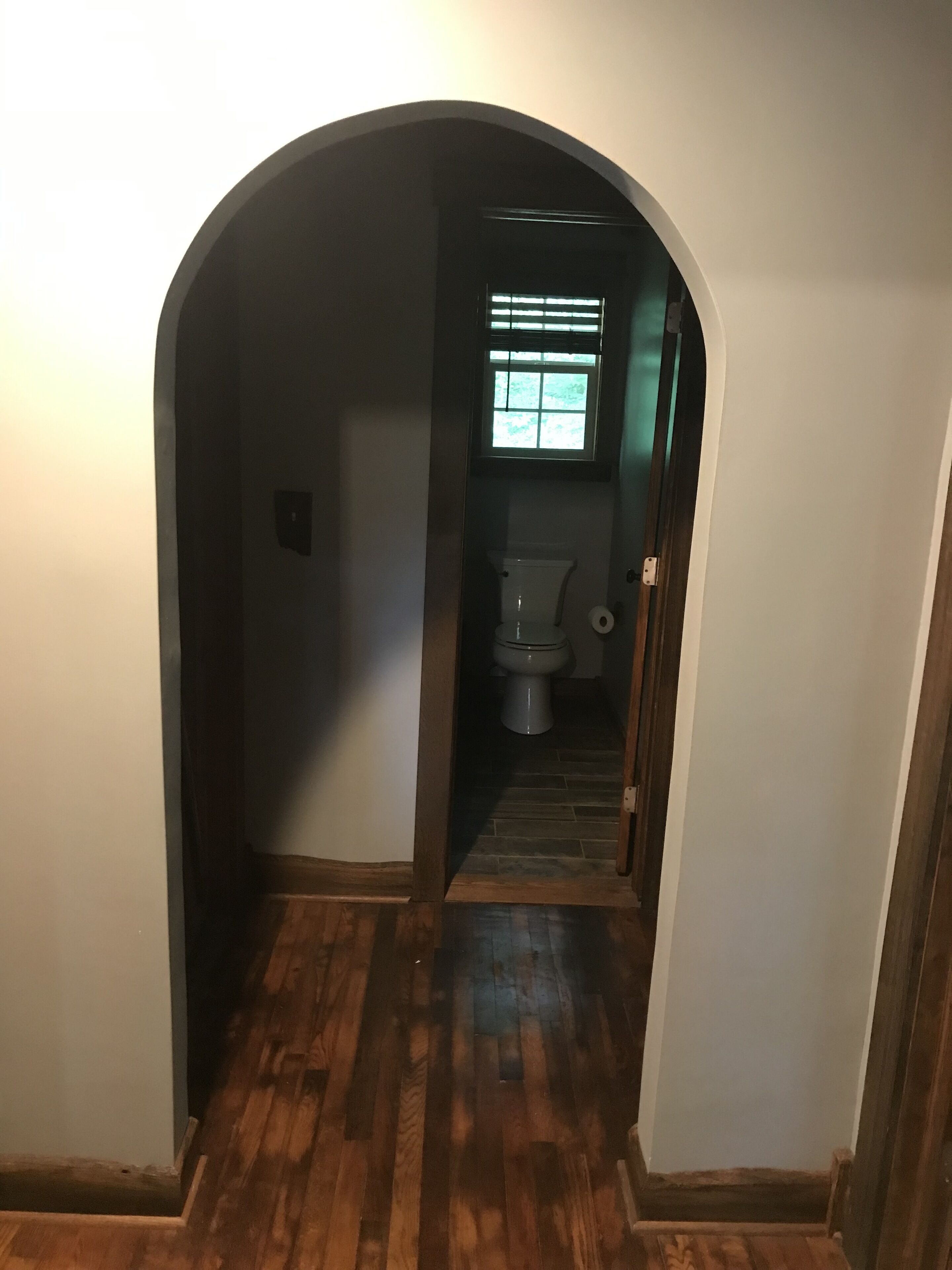 A narrow hallway with wooden flooring leading to a bathroom with a toilet and a window.