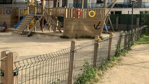 Un parque infantil con un tobogán y estructuras para escalar, rodeado por una valla y un edificio al fondo.