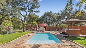 A rectangular swimming pool with stone borders, a hot tub, and a covered seating area in a backyard with trees and a wooden fence. - Villa 30 mins to Waco - Private Pool, Hot Tub, Gym and Fire Ring (Gatesville)