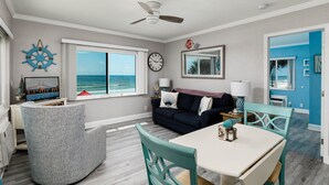 A living room with a view of the ocean, a ceiling fan, a clock, and a dining area with a table and chairs. - Cozy 1BR Condo w/ 0 min walk to beach! Spectacular views! Summer 2026 open! (North Redington Beach)