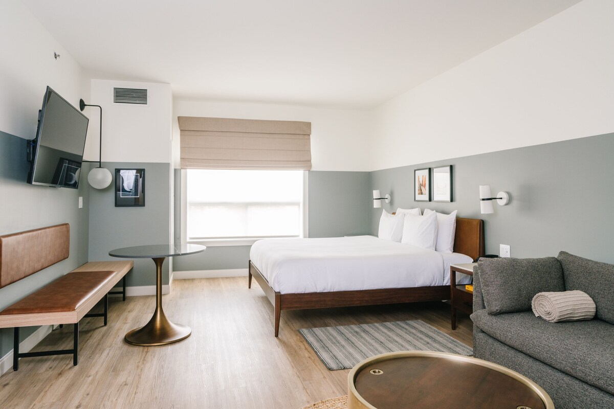 A modern hotel room with a bed, a sofa, a small table, a bench, and a TV mounted on the wall.