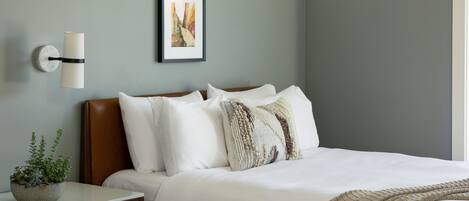 A hotel room with two beds, a bedside table with a plant, a framed picture on the wall, and a window with curtains.