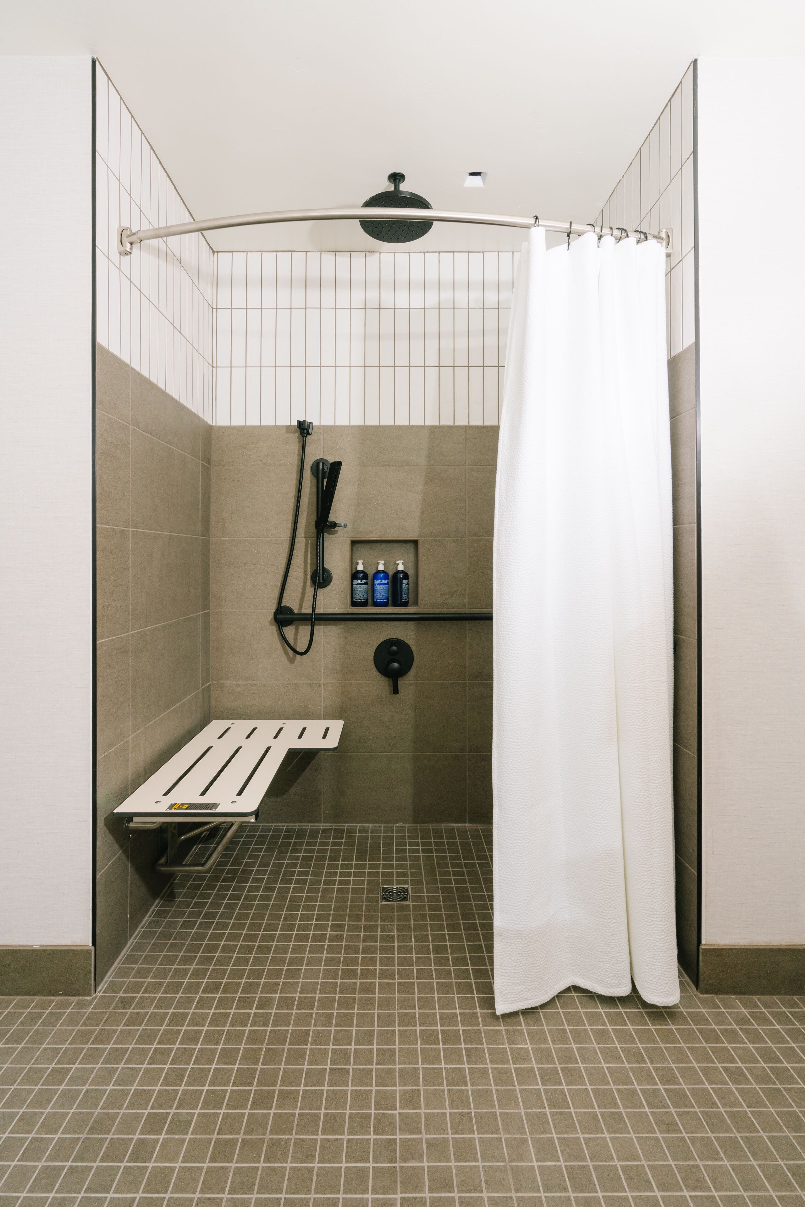 A shower stall with a white shower curtain, a handheld showerhead, a handheld shower head, and a shelf with toiletries.