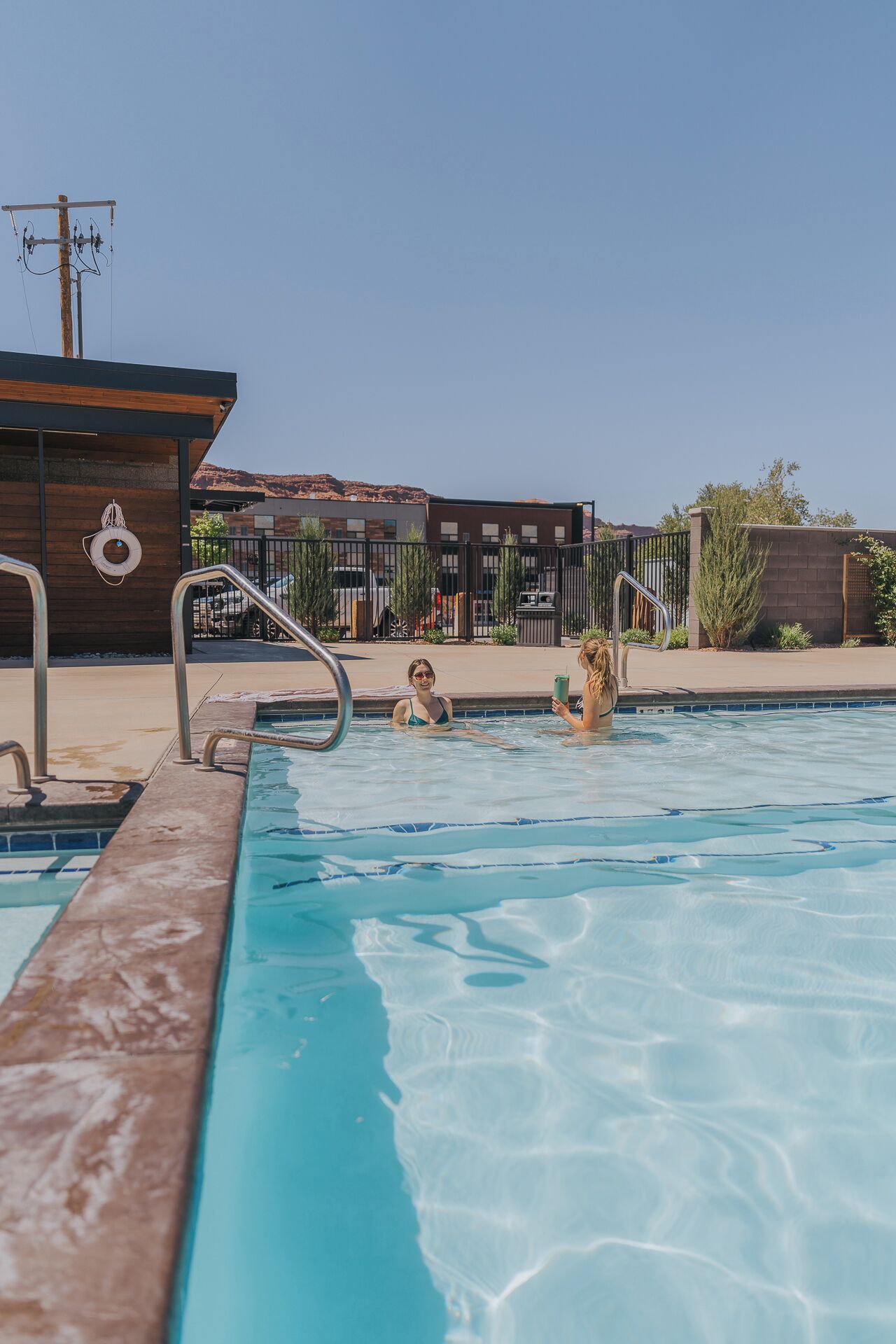 Una piscina con agua azul cristalina, barandales de metal y un flotador en el borde. En el fondo, un edificio con fachada de ladrillo y cielo despejado.