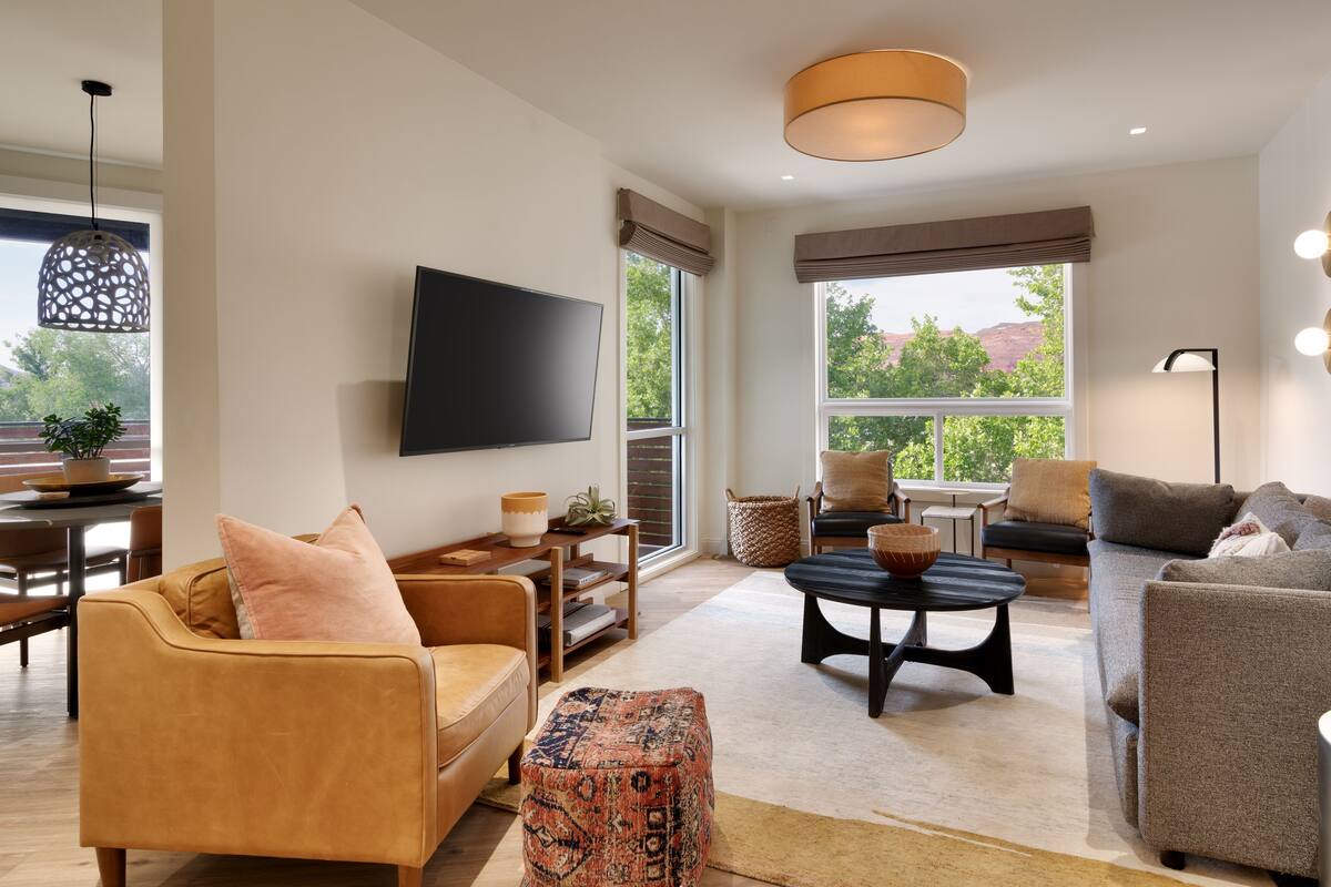 A modern living room with a sofa, armchair, and ottoman, a flat-screen TV, and a large window with a view of trees and hills.