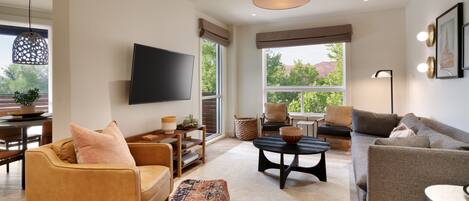 A modern living room with a sofa, armchair, and ottoman, a flat-screen TV, and a large window with a view of trees and hills.