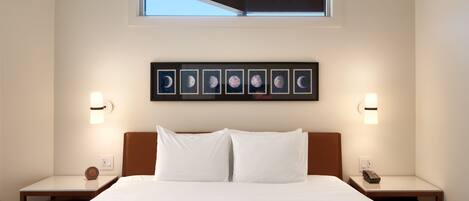 A hotel room with a large bed, two bedside tables, a nightstand with a phone, a wall-mounted clock, a framed picture of the moon phases, and a window with a view of the sky.