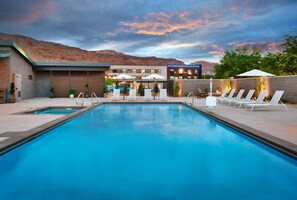 A rectangular swimming pool with lounge chairs and umbrellas, a building with a garage door, and a mountainous backdrop. - Gravity Haus Moab (Moab)