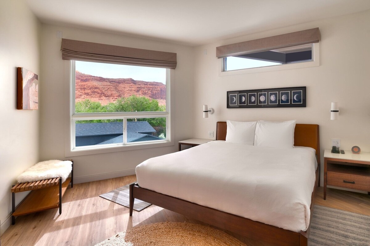 A bedroom with a large bed, a bench, a nightstand, a wall-mounted lamp, and a window with a view of red rock formations.