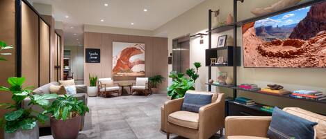 A modern hotel lobby with seating areas, a reception desk, and a large wall-mounted TV displaying a desert landscape.