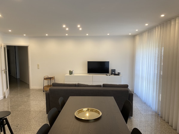 A modern living room with a flat-screen TV, a sofa, a dining table with a tray, and a large window with curtains.
